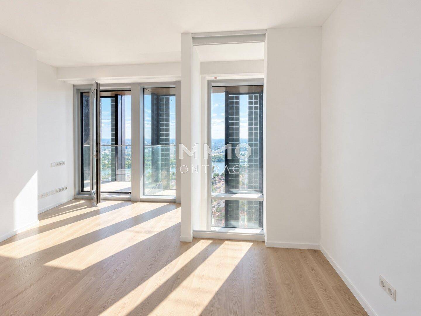 Heller Raum mit Parkettboden und großen Fenstern, die Zugang zu einem Balkon ermöglichen.