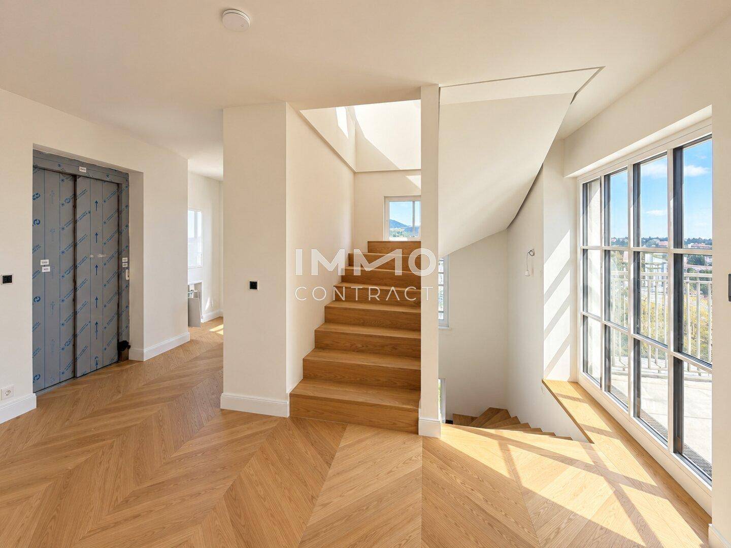 Heller Flur mit Holztreppe, Aufzug und großen Fenstern, die den Raum mit Licht füllen.
