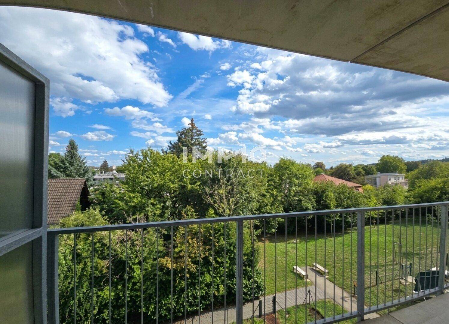 Weitläufiger Blick vom Balkon ins Grüne mit Bäumen, Rasenflächen und Sitzbänken unter blauem Himmel.