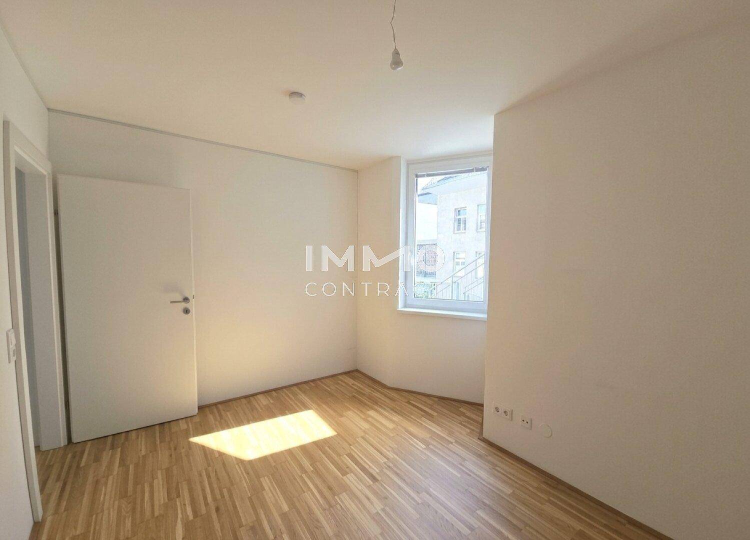Helles, unmöbliertes Zimmer mit Parkettboden und einem Fenster, das viel Sonnenlicht einfängt.