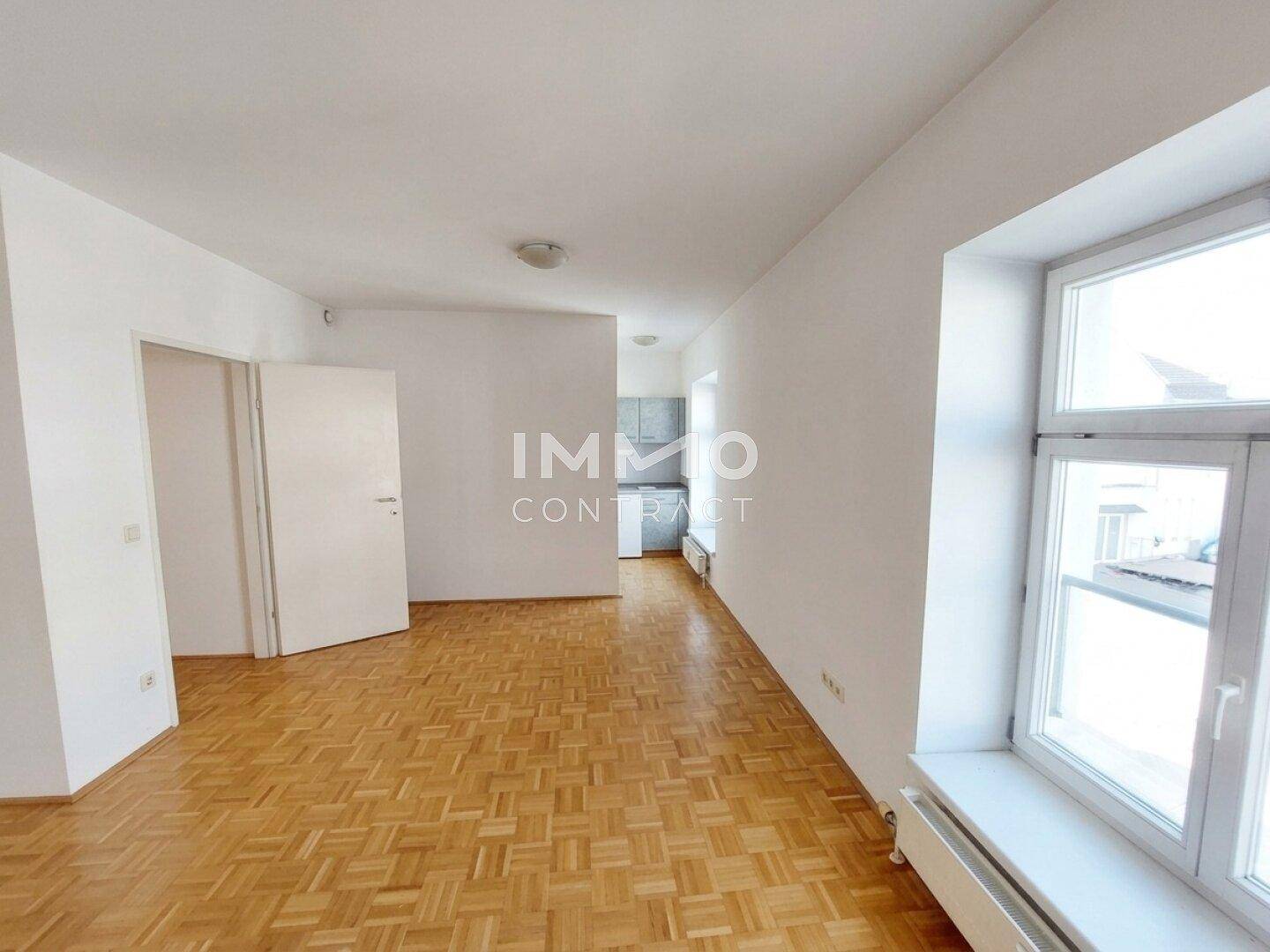 Leerer Raum mit Parkettboden und Fenster, der offen zur Küchenzeile führt.