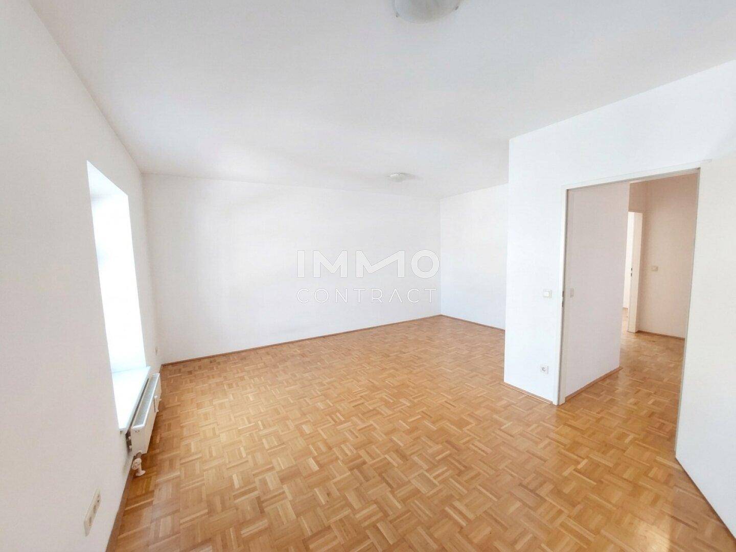 Geräumiges, leeres Wohnzimmer mit hellem Parkettboden und großen Fenstern für viel Tageslicht.
