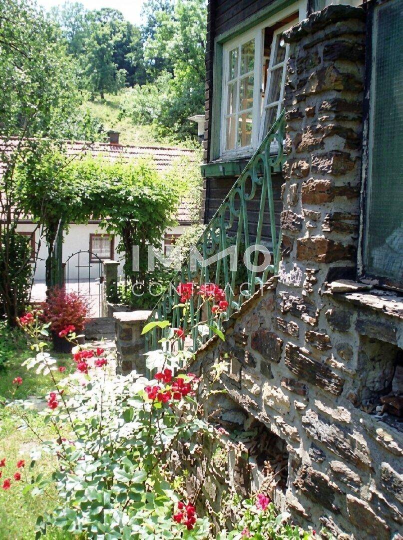 Detailansicht des Hauses mit Steinfundament, grünem Geländer und blühenden Rosen im Garten.