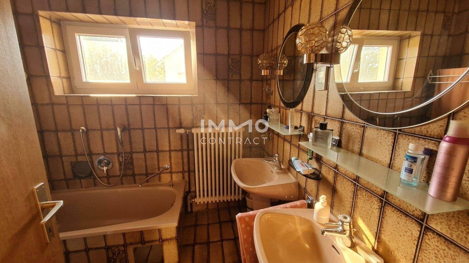Badezimmer im 80er-Jahre-Stil mit Badewanne, Doppelwaschbecken und zwei Fenstern.