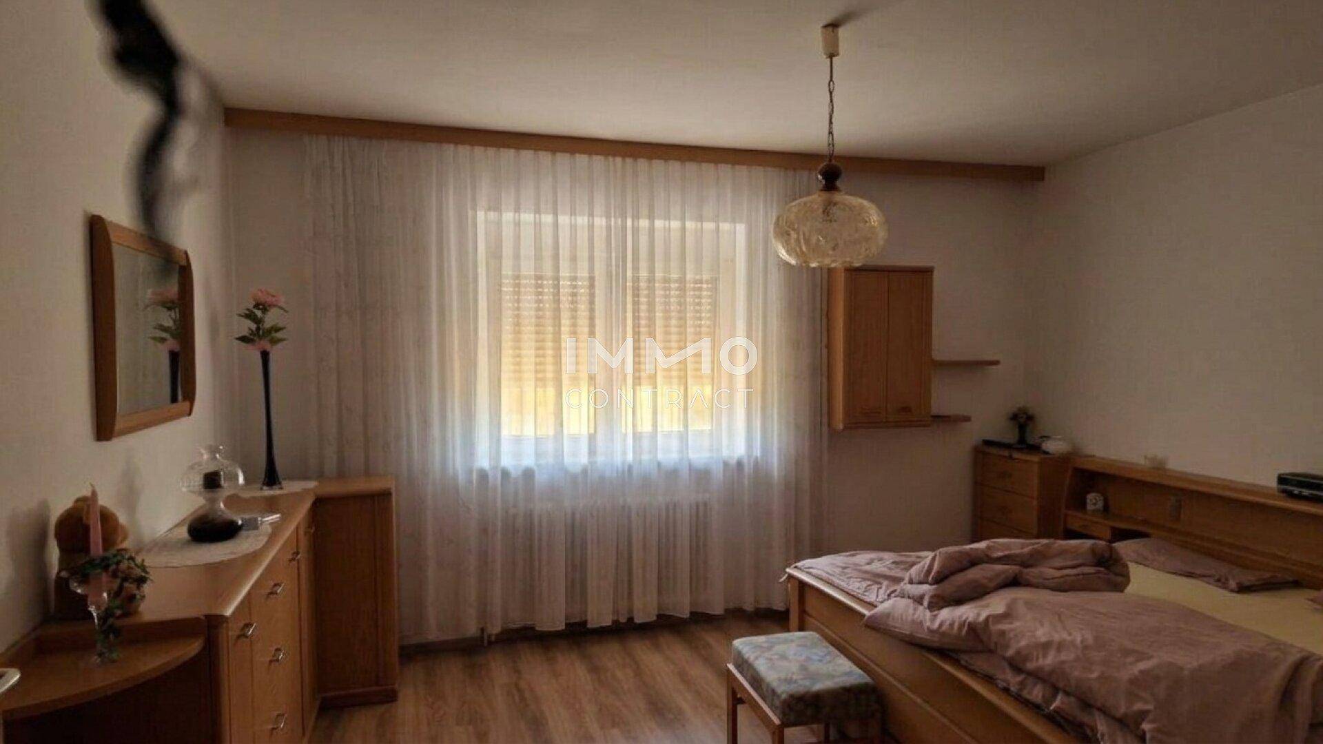 Schlafzimmer mit Doppelbett, Holzmöbeln und großem Fenster mit Vorhängen.