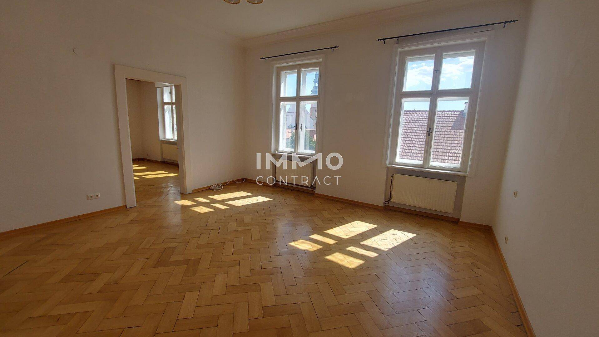 Geräumiges Zimmer mit Fischgrätparkett und großen Fenstern, die viel Tageslicht hereinlassen.