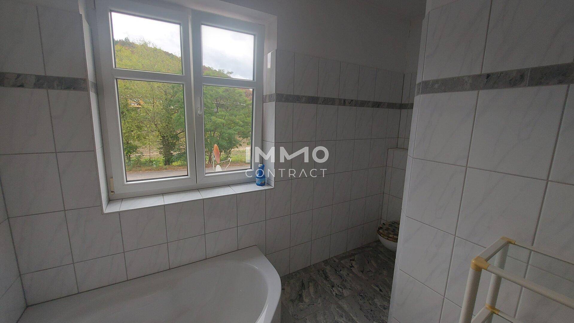 Badezimmer mit Fliesen, Badewanne und Fenster, das einen Blick ins Grüne ermöglicht.
