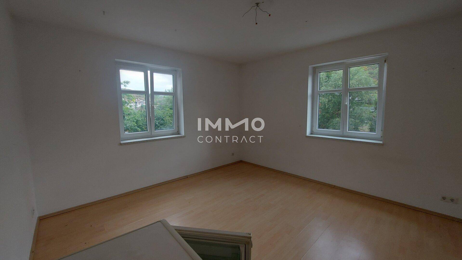 Helles Schlafzimmer mit Holzboden und zwei Fenstern, die einen Blick ins Grüne bieten.