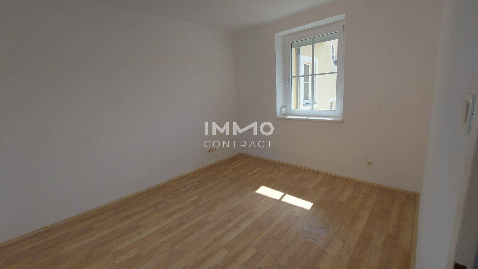Helles, leeres Zimmer mit Parkettboden und einem großen Fenster, ideal als Schlafzimmer.