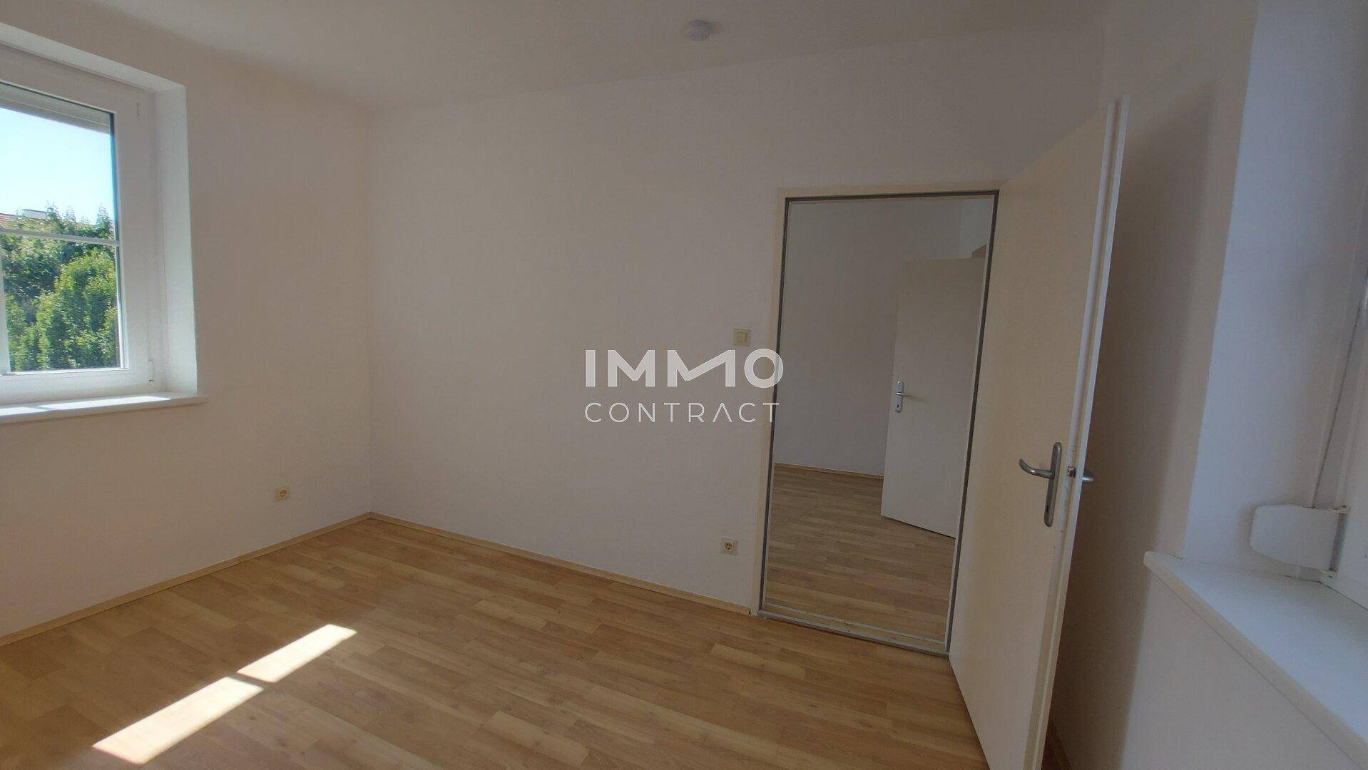 Geräumiges Zimmer mit Parkettboden, Fenster und einer Tür mit integriertem Spiegel.
