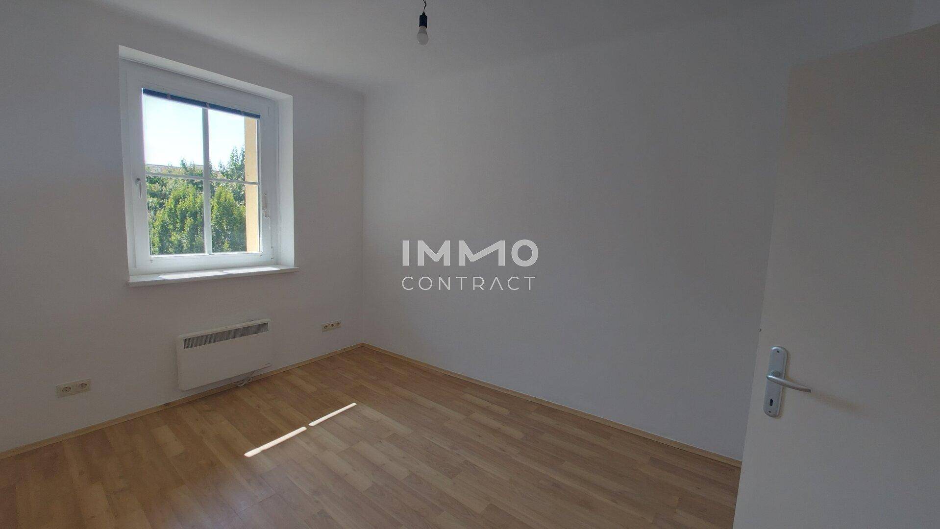 Lichtdurchflutetes Zimmer mit Parkettboden, weißen Wänden und einem Heizkörper unter dem Fenster.