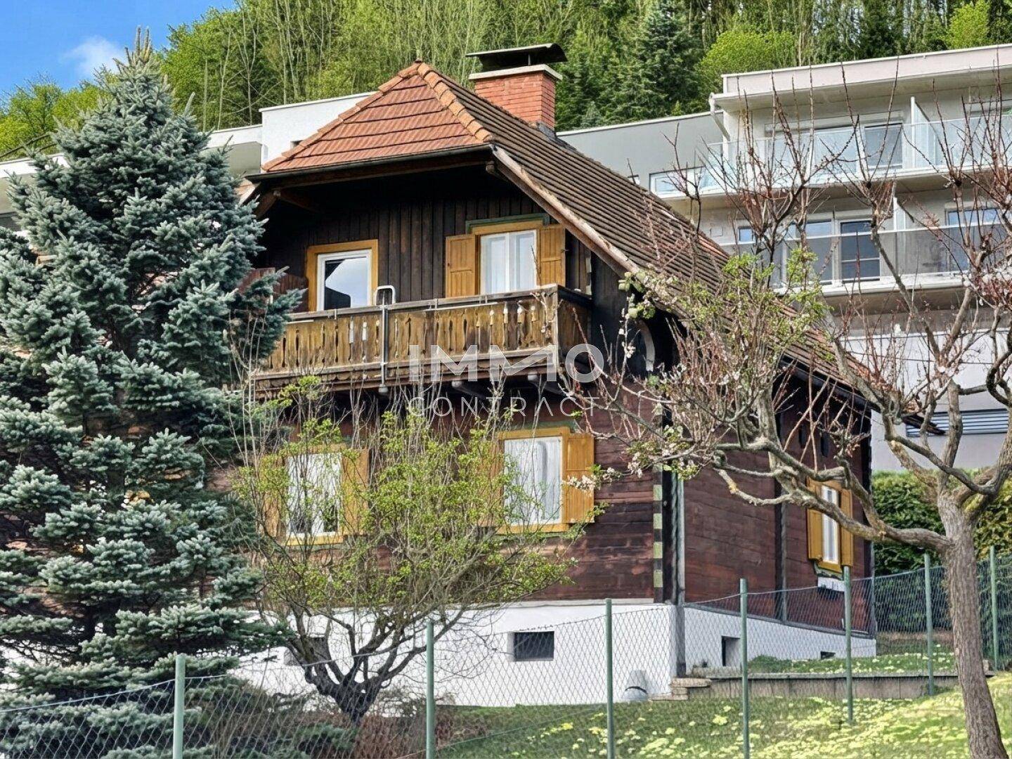 Traditionelles Holzhaus im Chalet-Stil mit Balkon, Ziegeldach und grüner Umgebung, im Hintergrund ein modernes Gebäude.