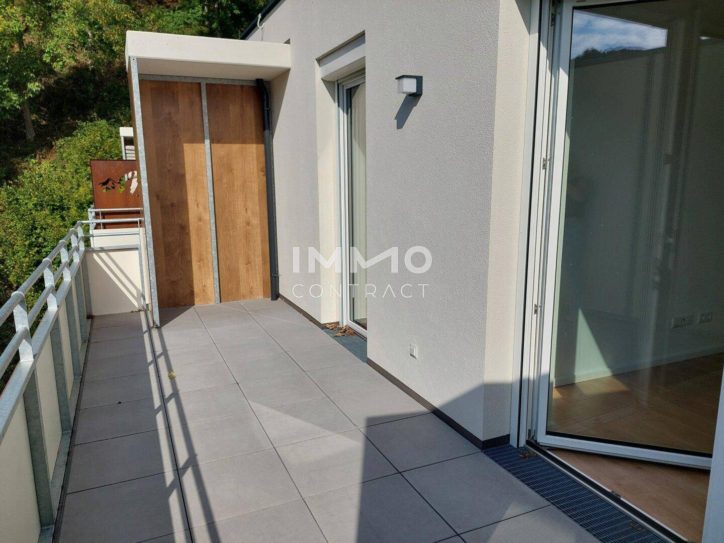 Moderner Balkon mit Fliesenboden und Metallgeländer, umgeben von grüner Natur und Bäumen.