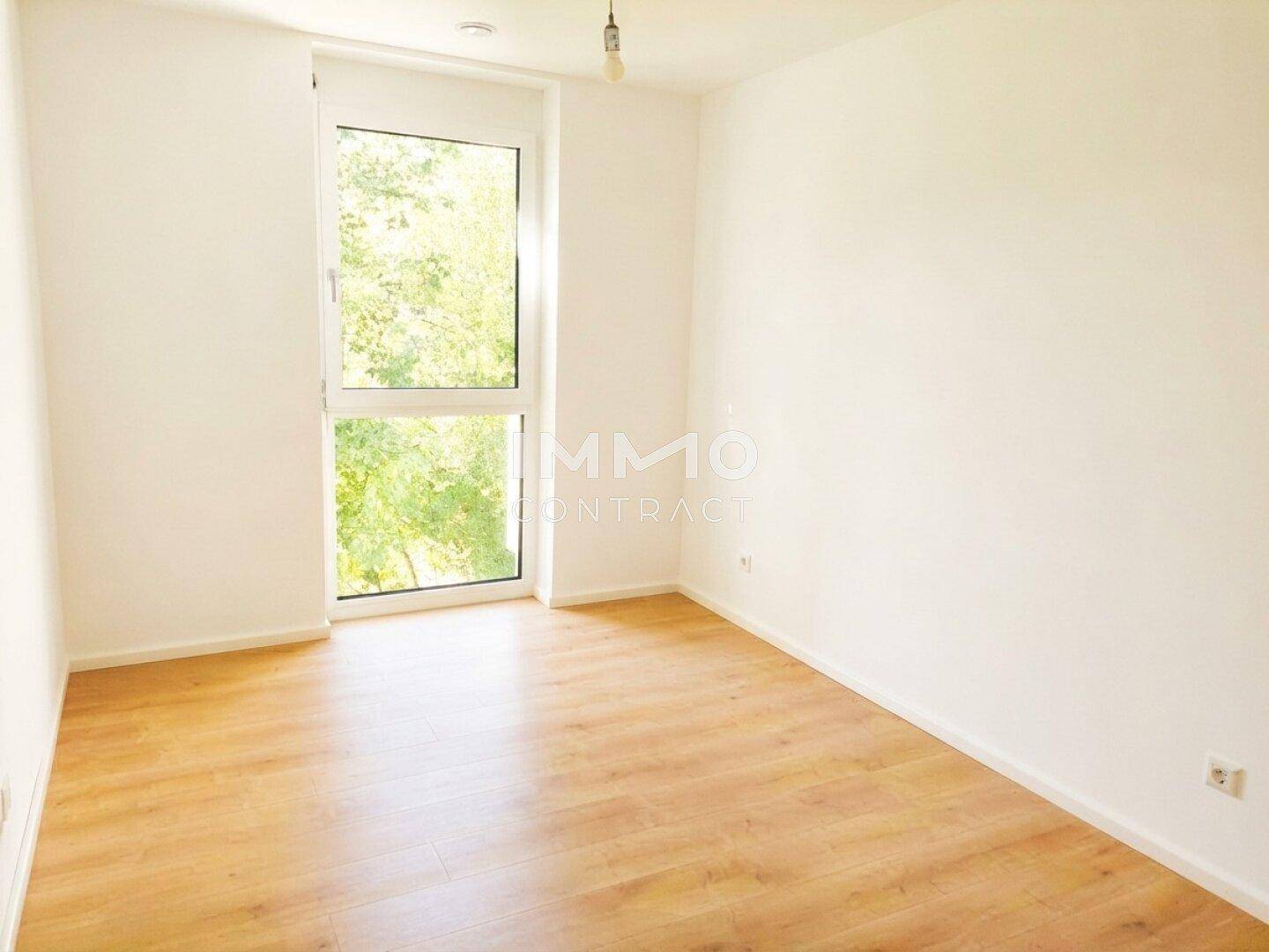 Leeres Schlafzimmer mit Laminatboden und großem Fenster, das viel Tageslicht hereinlässt.