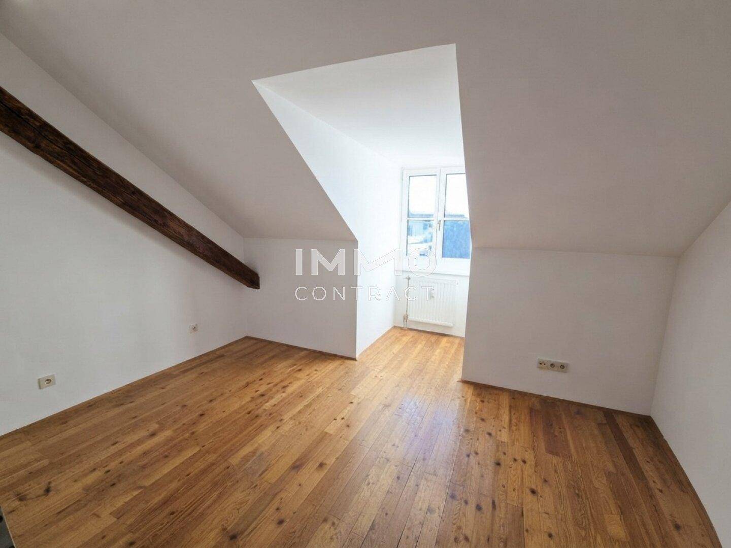 Geräumiger Dachgeschossraum mit Holzboden, weißen Wänden und einem sichtbaren Holzbalken.
