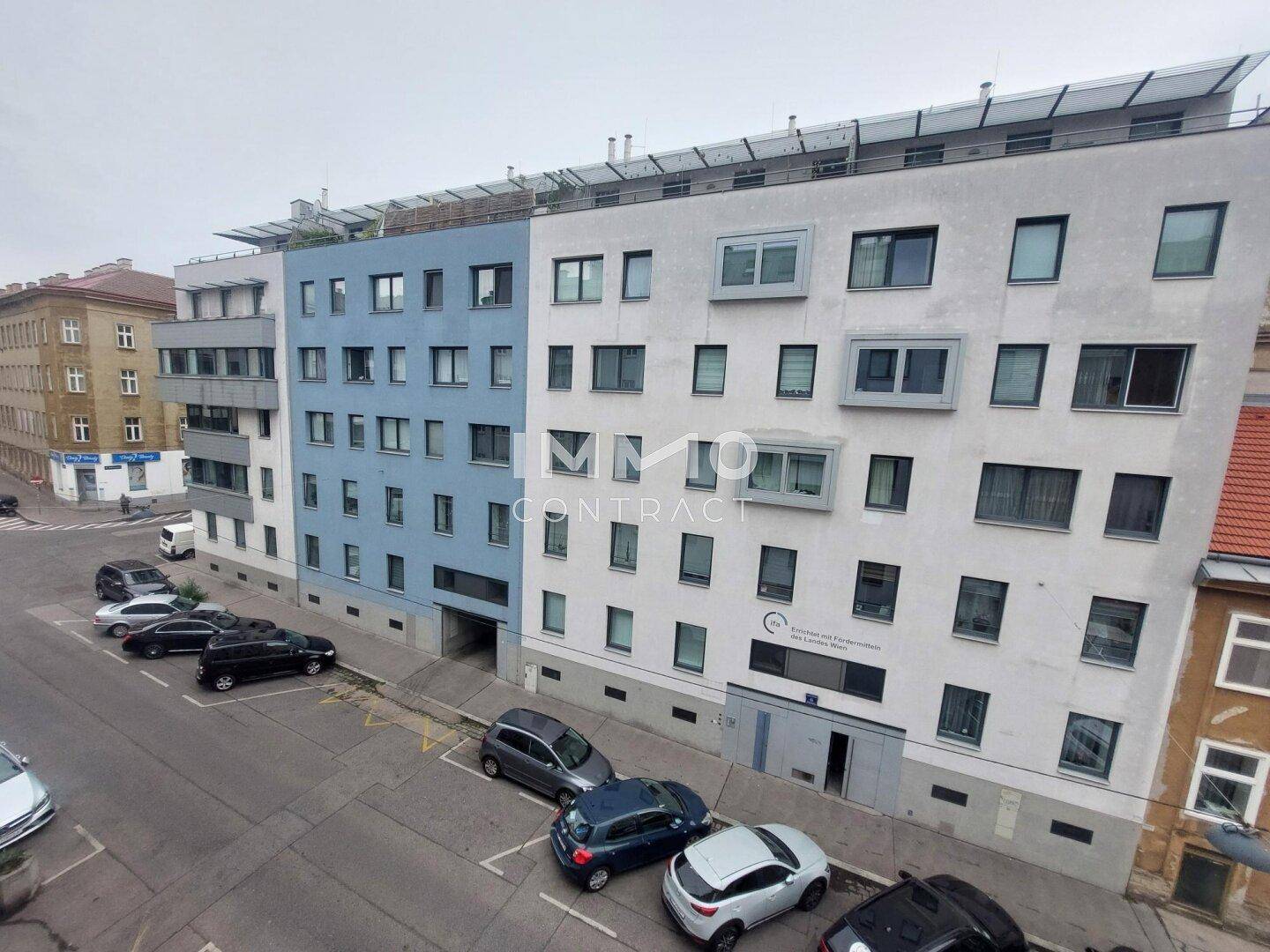 Außenansicht des Gebäudes mit moderner Fassade und parkenden Fahrzeugen auf der Straße.