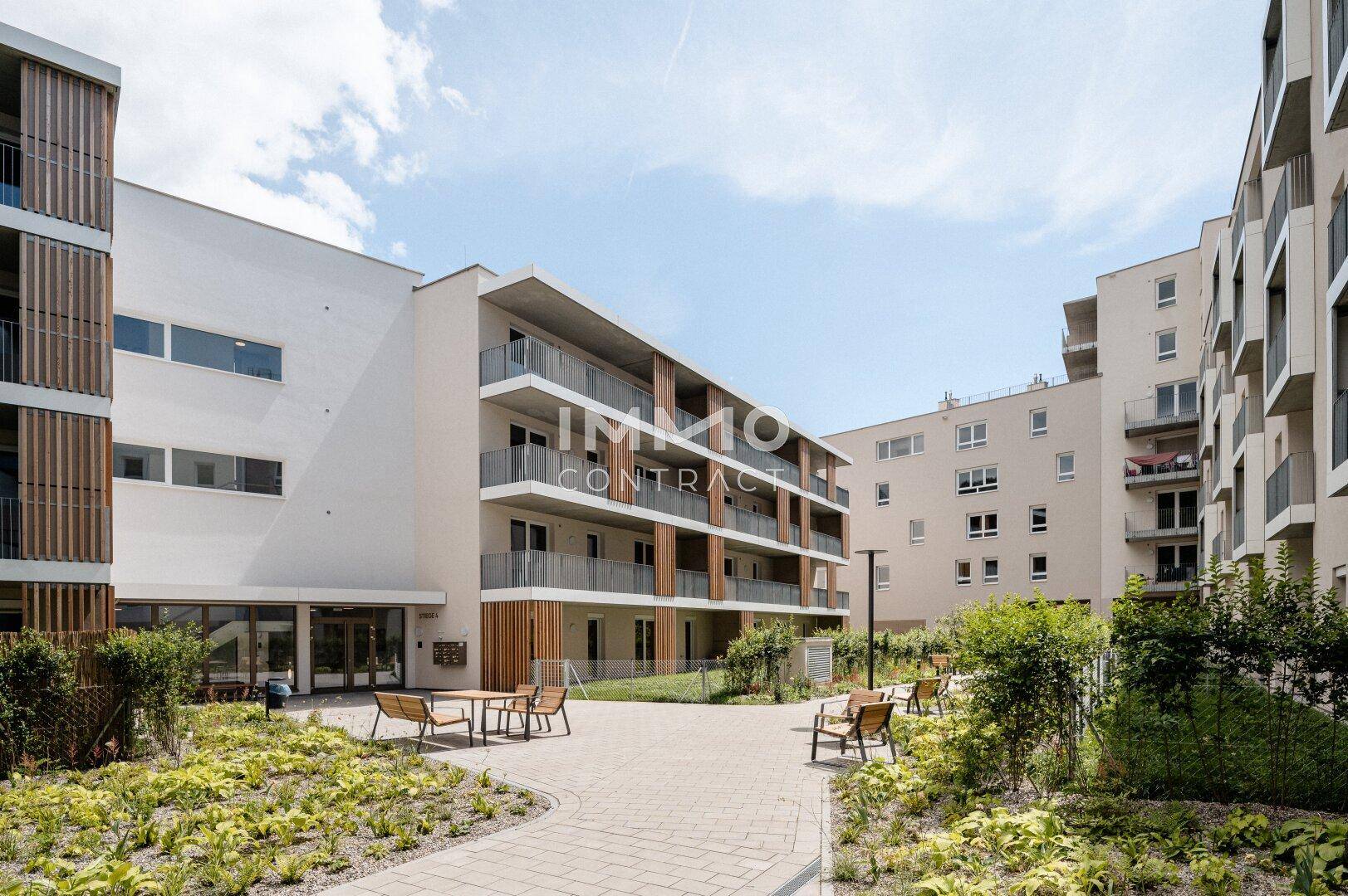 Ansicht des modernen Apartmentkomplexes mit begrüntem Innenhof und weiteren Sitzgelegenheiten im Freien.