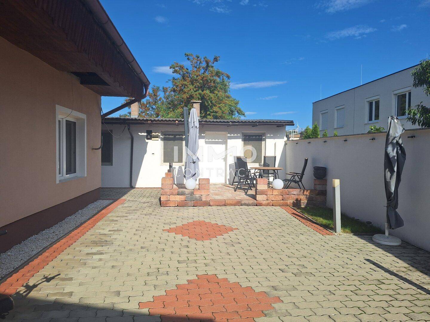 Sonniger gepflasterter Außenbereich mit Sitzgelegenheiten und einem Sonnenschirm vor dem Gebäude.