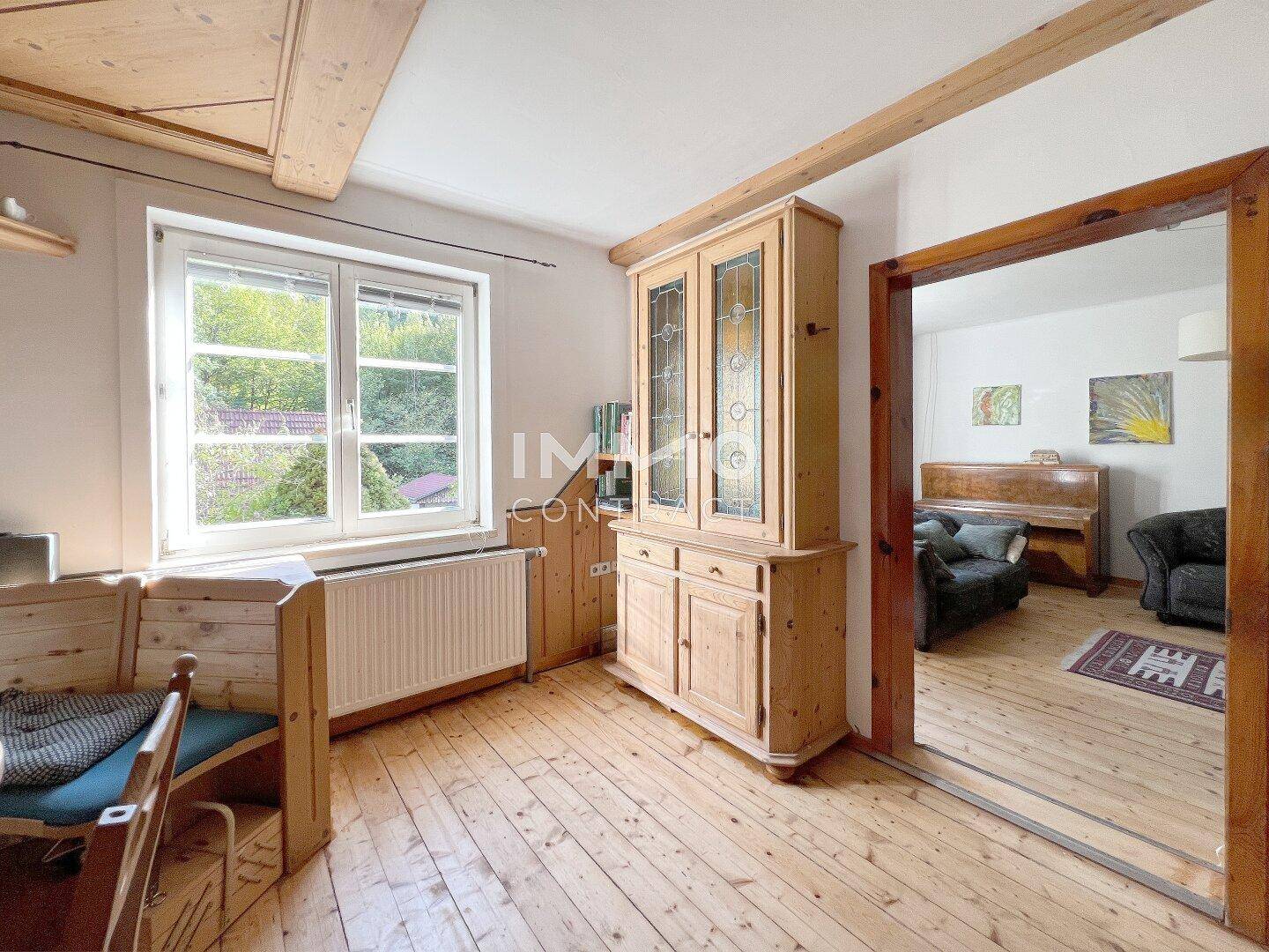 Heller Essbereich mit Holzboden und großem Fenster, angrenzend an ein weiteres Wohnzimmer mit Klavier.