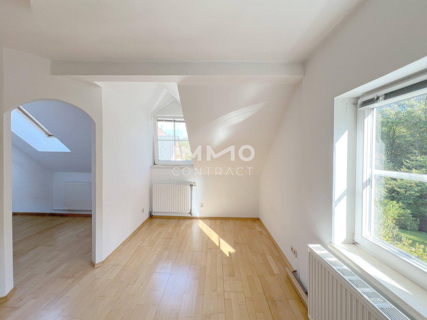 Helles Dachzimmer mit schrägen Wänden, Holzfußboden und einem Fenster mit Blick ins Grüne.
