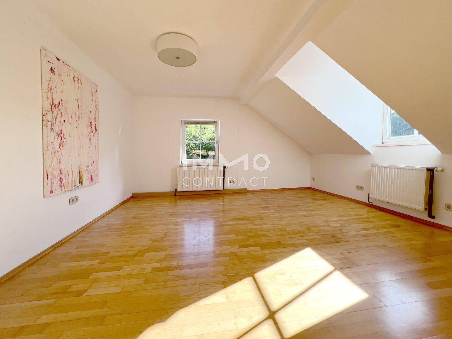 Helles und geräumiges Dachzimmer mit Holzboden, schrägen Wänden und zwei Fenstern.