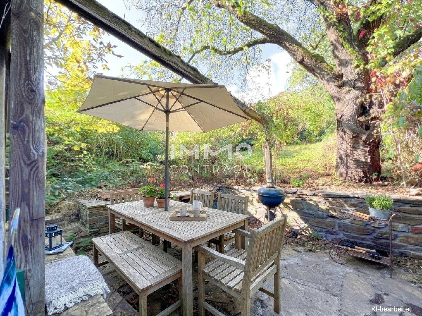 Gemütlicher Sitzbereich im Garten mit Holztisch, Sonnenschirm und Grillplatz, umgeben von Natur.