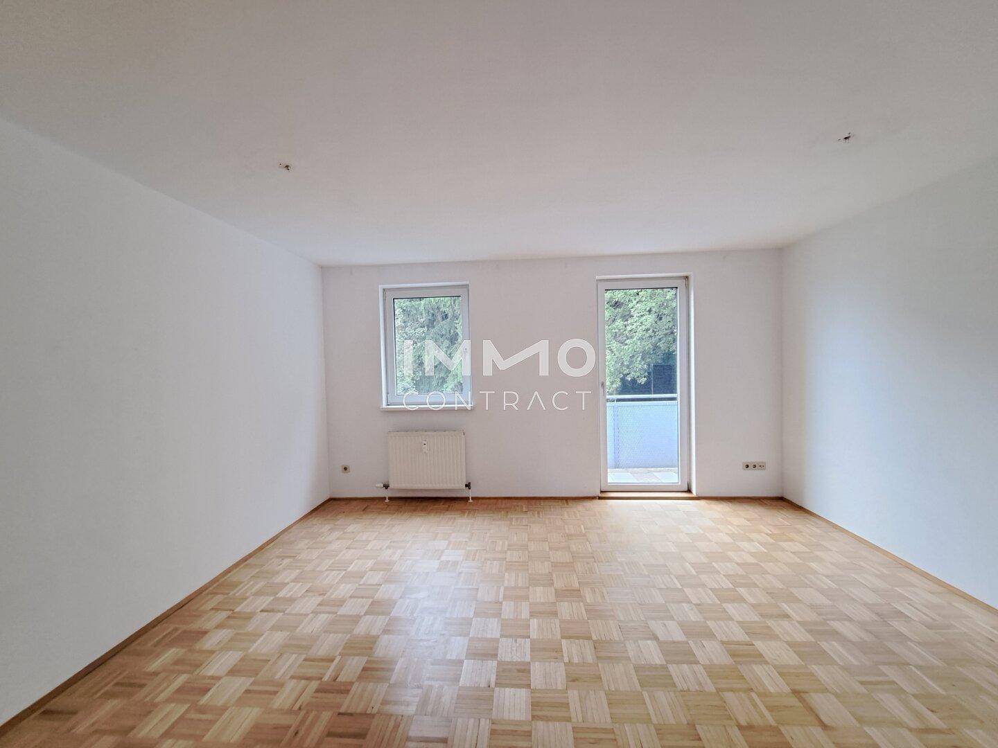 Helles, leeres Wohnzimmer mit Fischgrätparkettboden und Zugang zu einem Balkon.