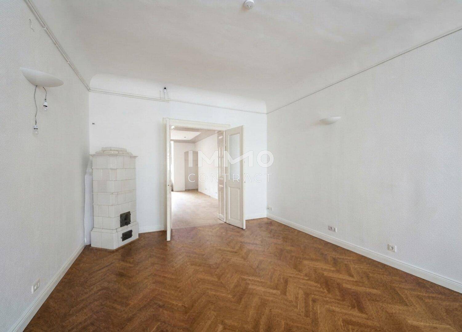 Leerer Raum mit Fischgrätparkett, weißem Kachelofen und Doppeltüren zu einem weiteren Zimmer.