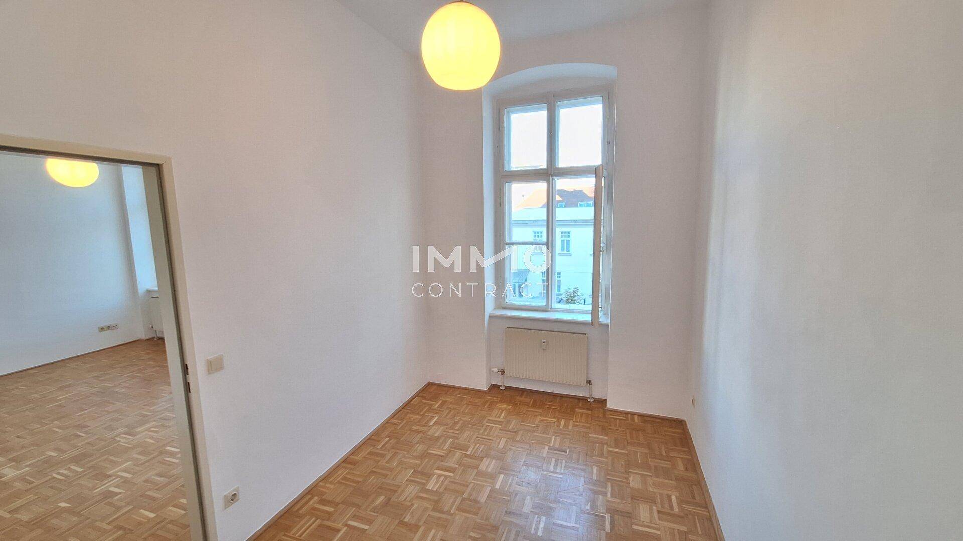 Helles Schlafzimmer mit Parkettboden und einem hohen Fenster, das viel Tageslicht hereinlässt.