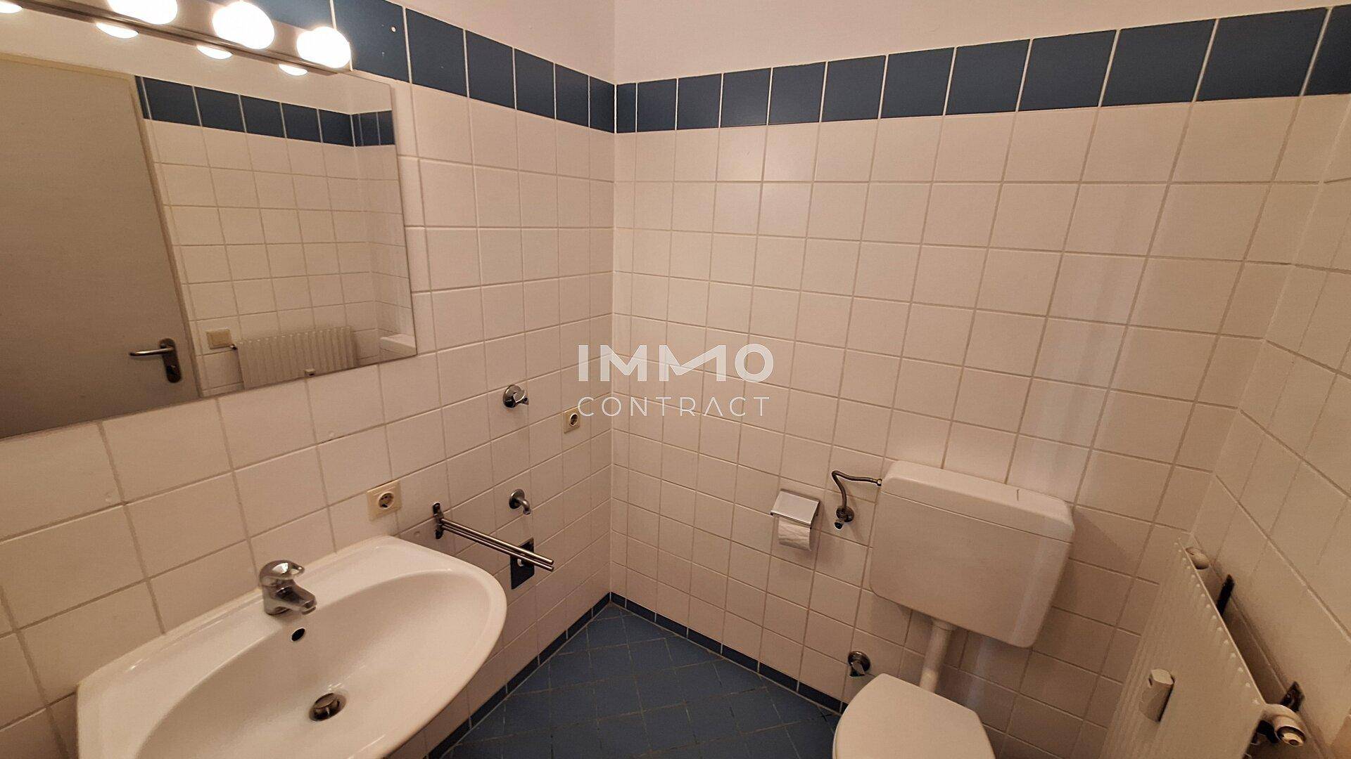 Einfaches WC mit weiß-blauen Fliesen, Waschbecken und Spiegel, funktional eingerichtet.