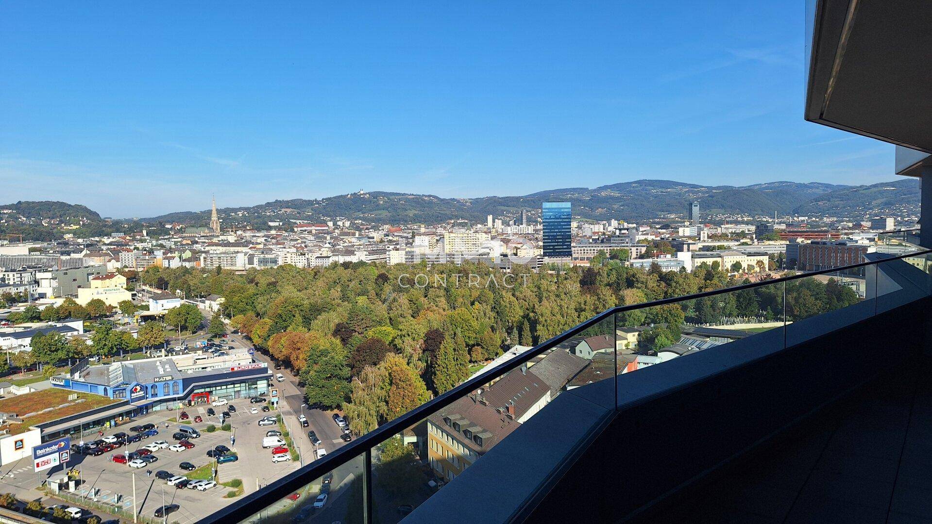 Weitläufiger Stadtblick von einem Balkon mit Glasbrüstung unter blauem Himmel.