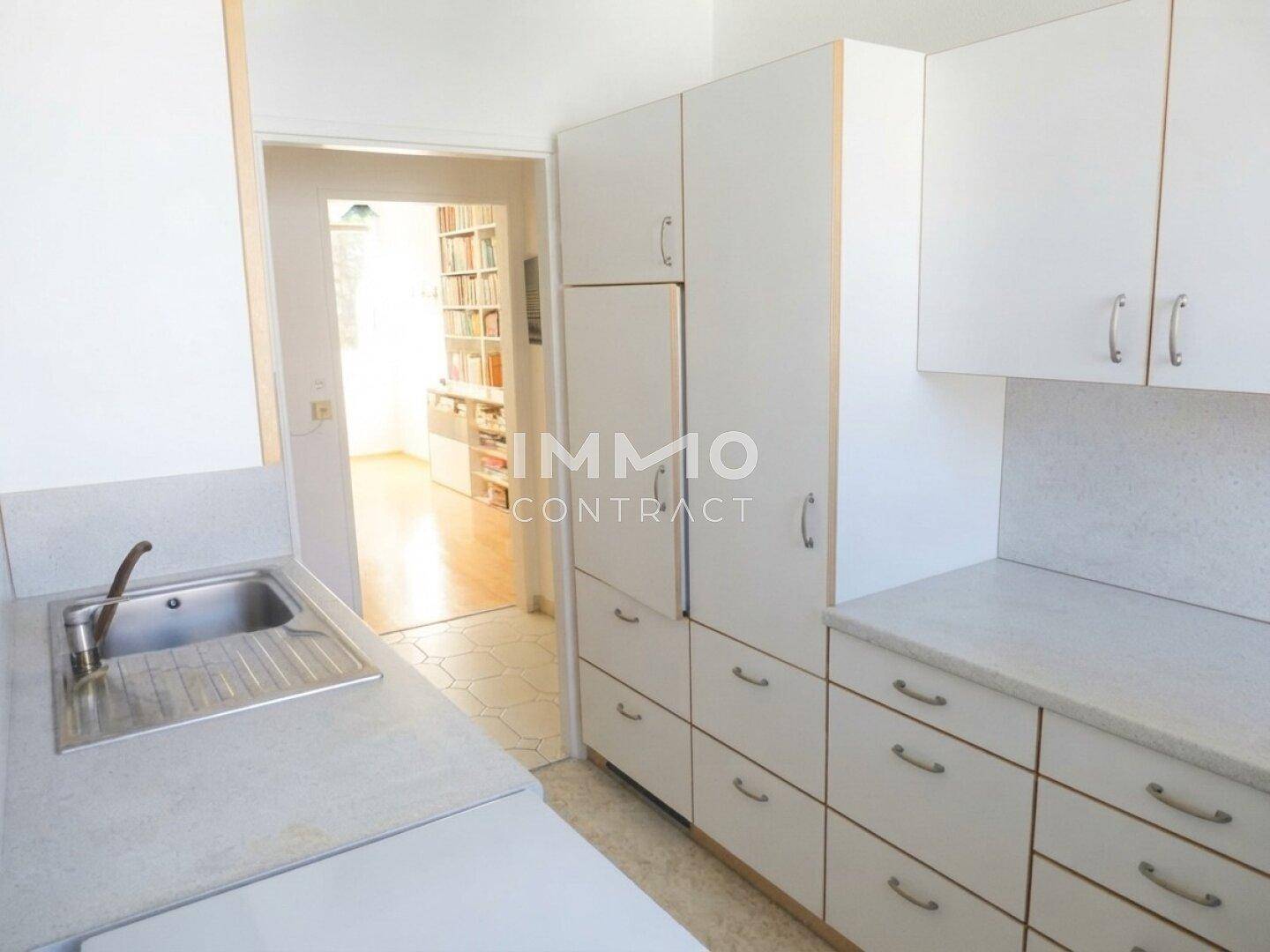 Einfache Küche mit weißen Schränken, Spüle und Kühlschrank, die Zugang zu einem weiteren Raum bietet.