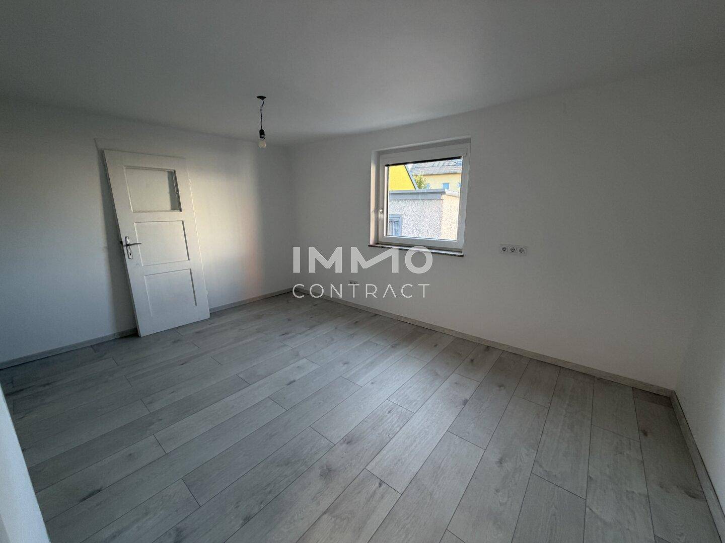 Leeres Zimmer mit hellem Laminatboden und einem Fenster mit Blick ins Grüne.