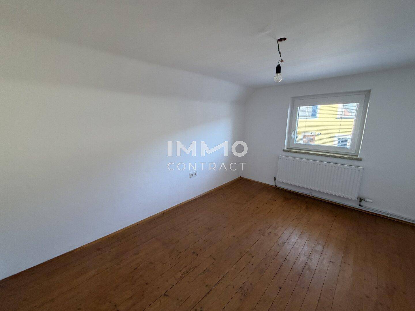 Leeres Zimmer mit Holzdielenboden, weiß gestrichenen Wänden und Heizkörper unter dem Fenster.