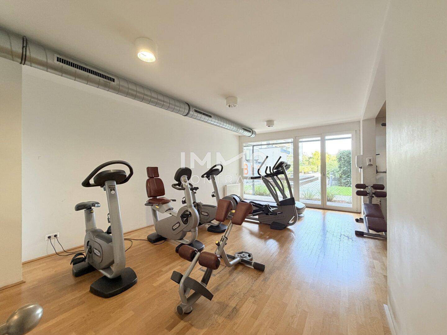 Geräumiger Fitnessraum mit verschiedenen Trainingsgeräten und großen Fenstern mit Blick ins Grüne.