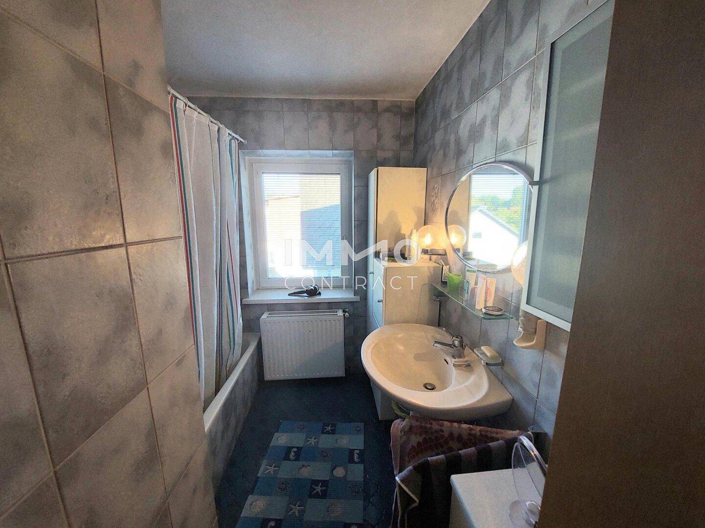 Badezimmer mit grauen Fliesen, Badewanne mit Duschvorhang und Fenster.