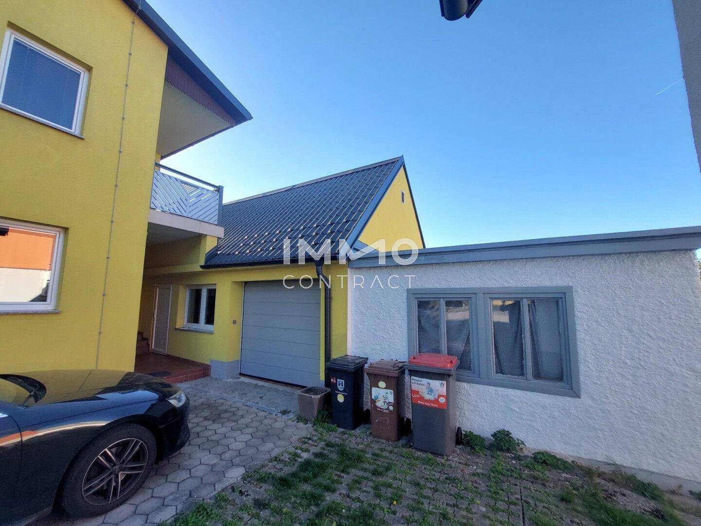 Seitenansicht des gelben Hauses mit Garage, Balkon und gepflasterter Einfahrt.