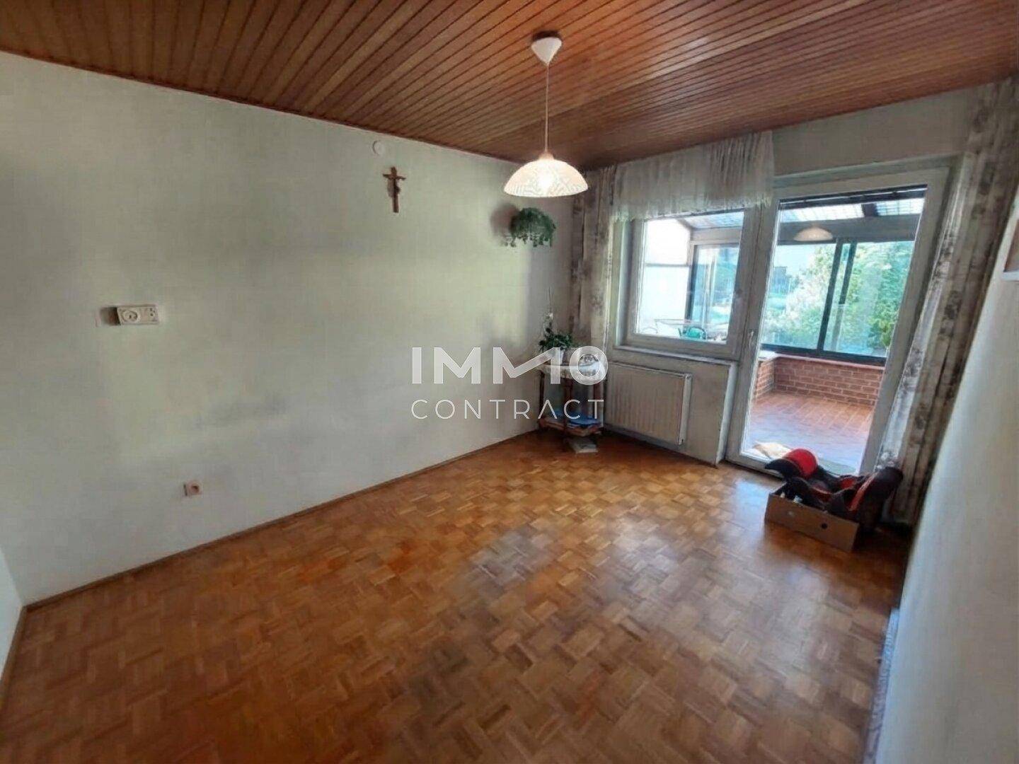 Wohnzimmer mit Parkettboden, Holzdecke und Zugang zu einem verglasten Balkon.