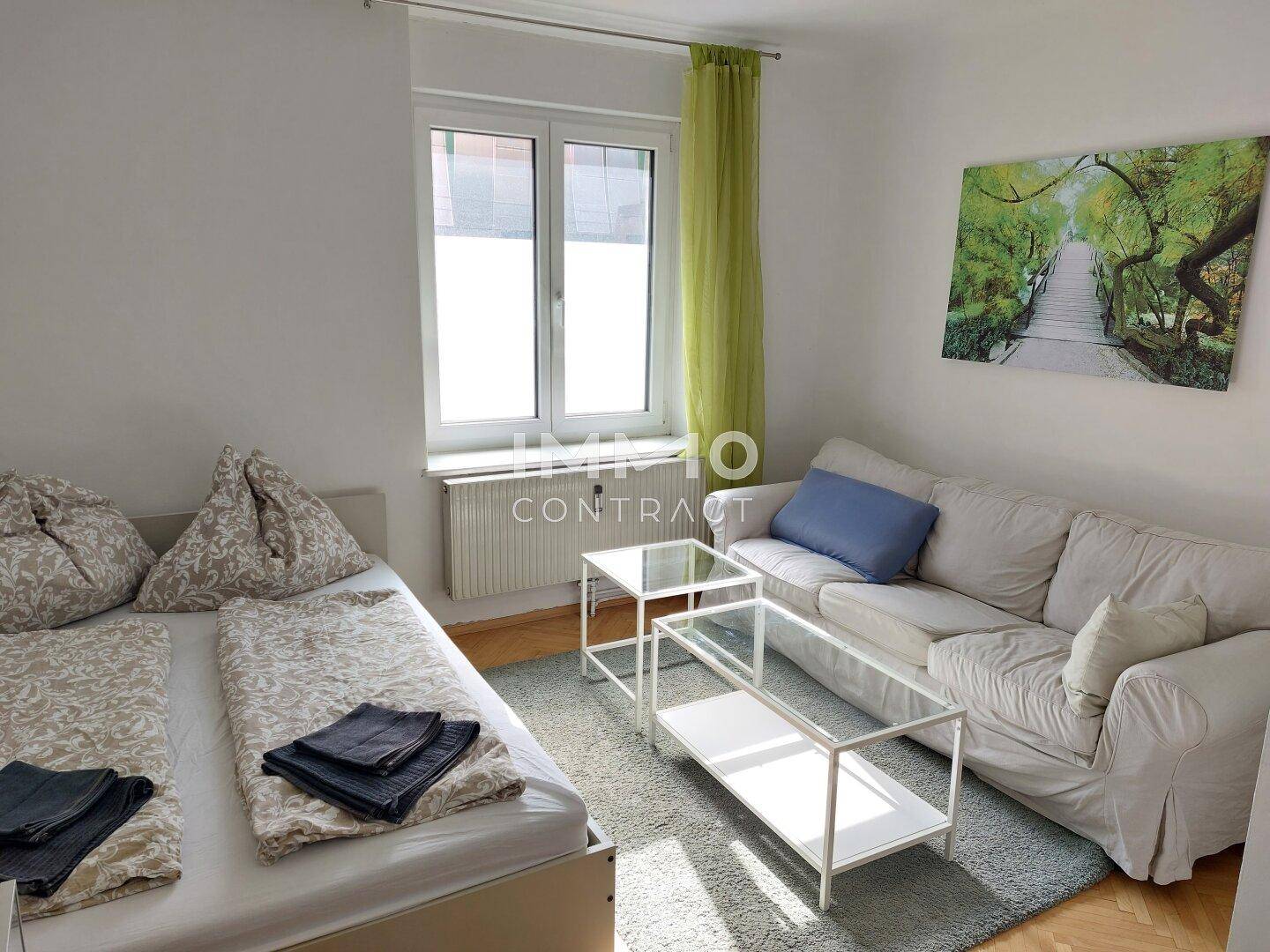 Helles Wohnzimmer mit Schlafsofa, Teppichboden und Blick ins Grüne durch das Fenster.