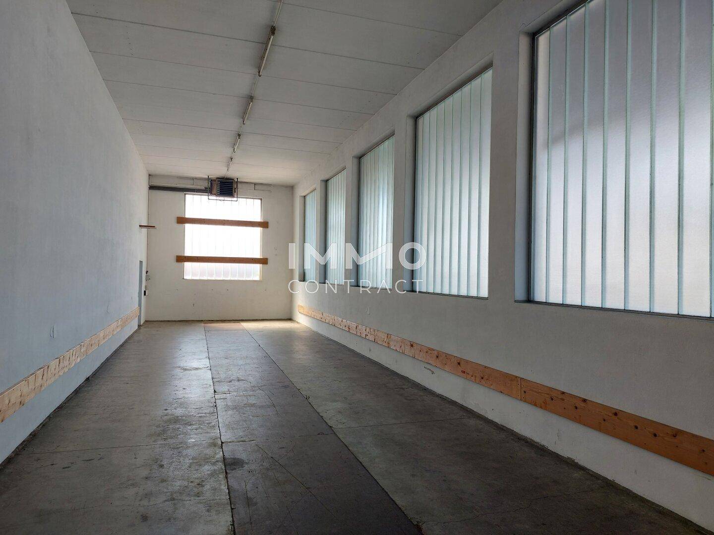 Langer, leerer Raum mit Betonboden und hohen Fenstern, geeignet als Lager- oder Arbeitsbereich.