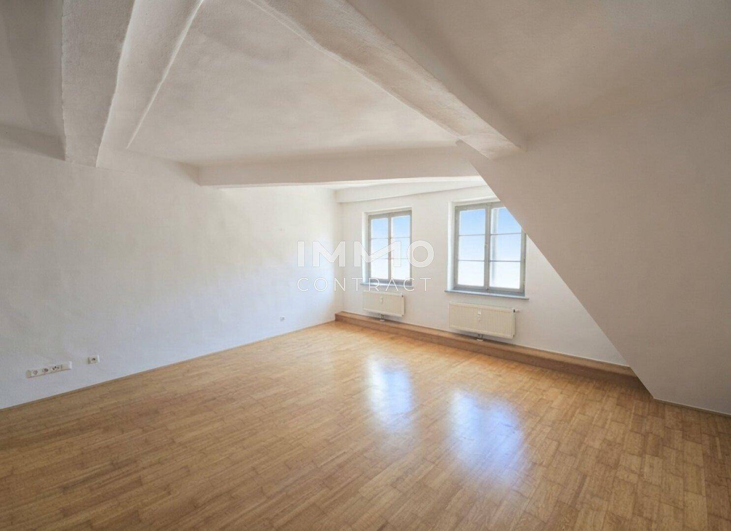 Heller Dachbodenraum mit Holzboden, weißen Wänden und zwei Fenstern, die viel Tageslicht hereinlassen.