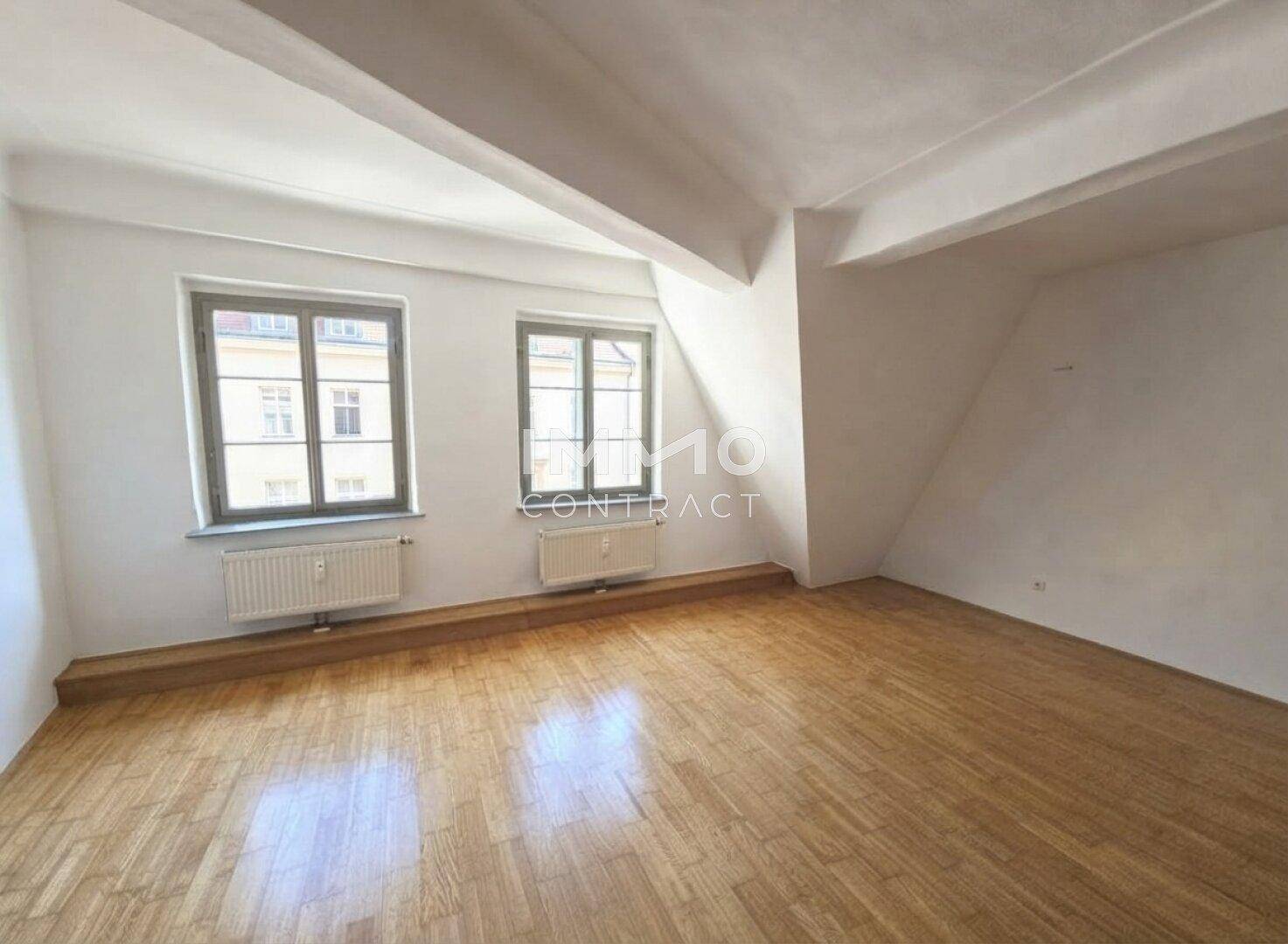 Geräumiger Dachbodenraum mit Parkettboden, weißen Wänden und zwei Fenstern, die den Raum erhellen.