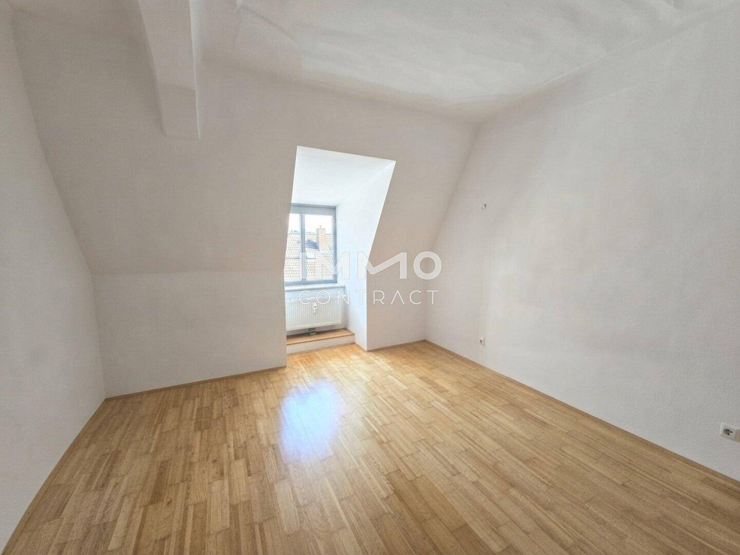 Heller Raum im Dachgeschoss mit Holzboden, weißen Wänden und einem einzelnen Fenster, das Tageslicht spendet.