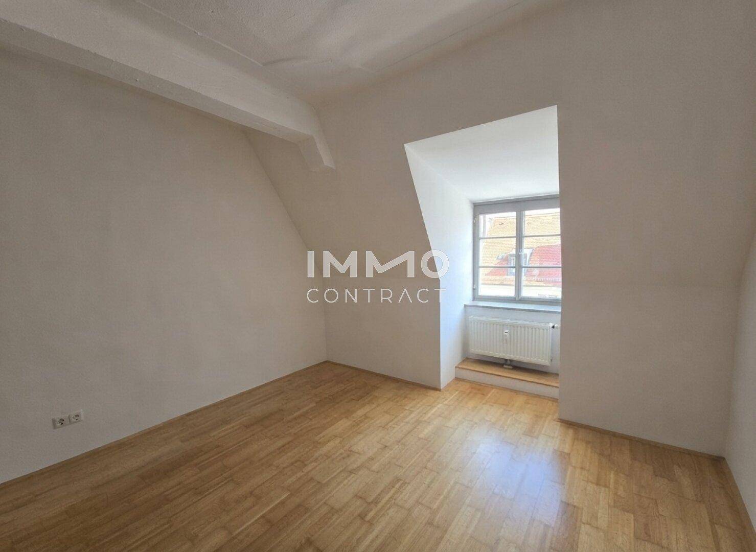 Gemütlicher Dachbodenraum mit Parkettboden, weißen Wänden und einem Fenster, das den Raum mit Licht füllt.