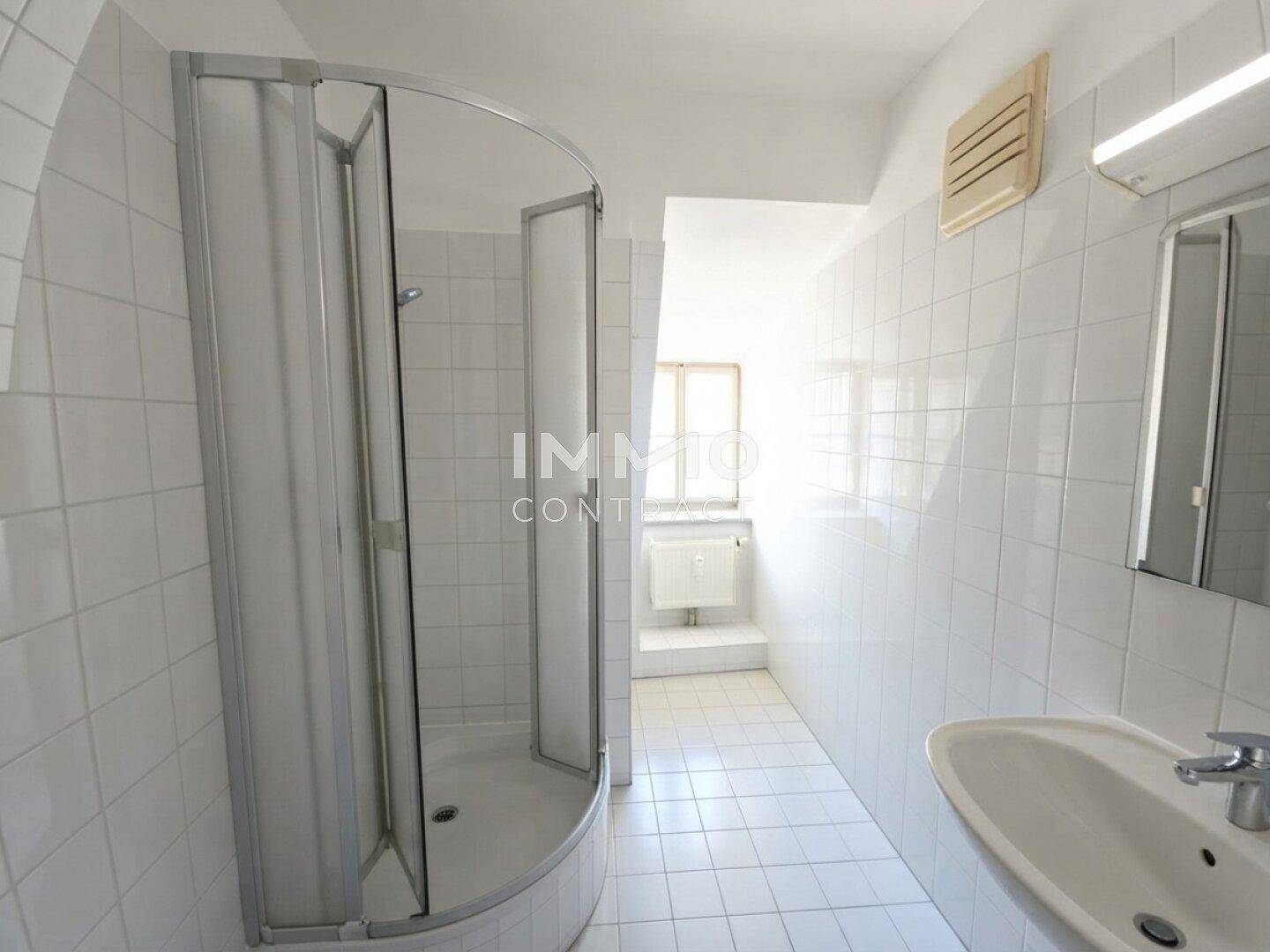 Badezimmer mit weißen Fliesen, einer Eckdusche, Waschbecken und einem Fenster für natürliche Belüftung.
