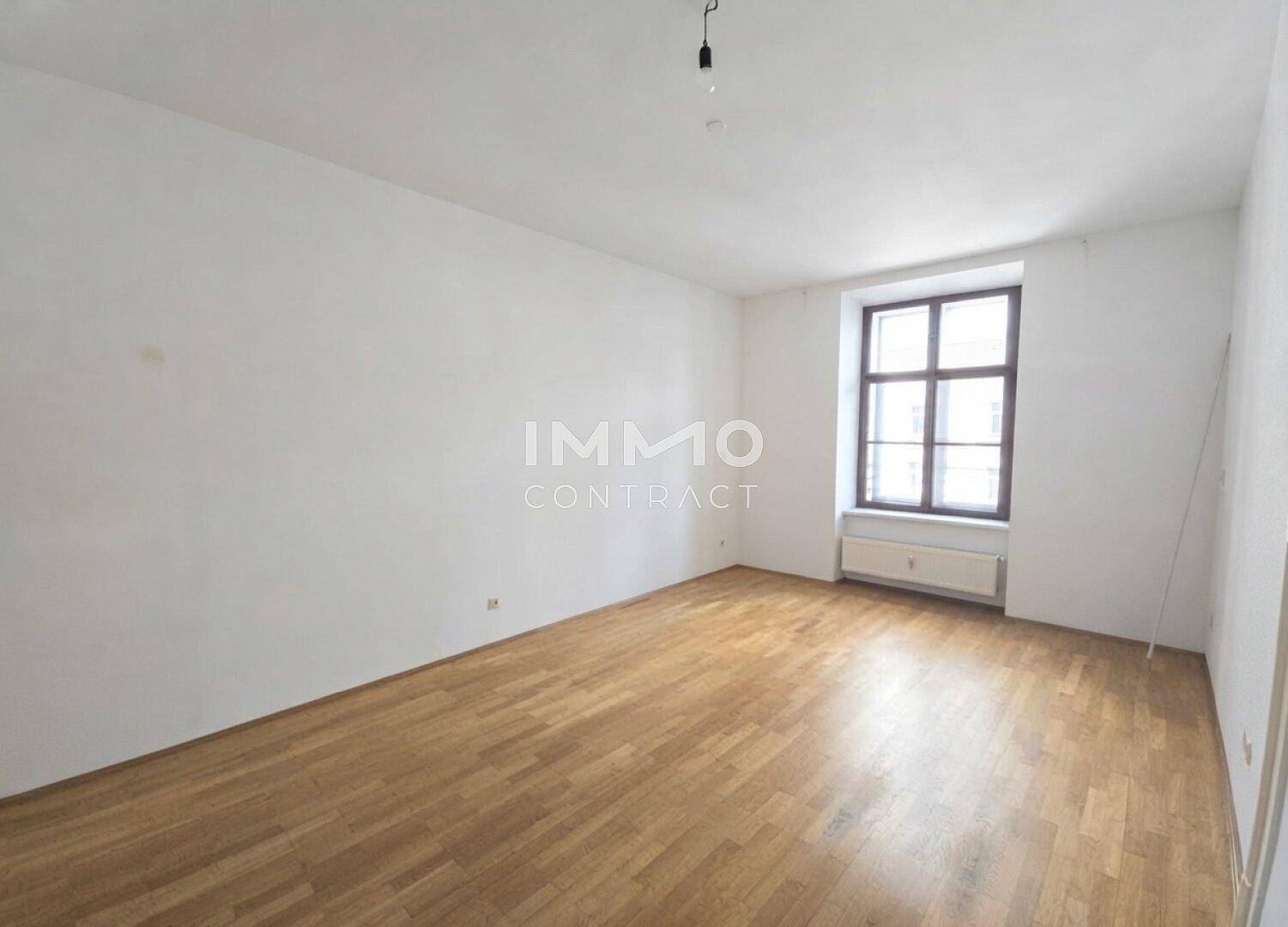 Heller, leerer Raum mit Parkettboden und einem Fenster, das viel Tageslicht hereinlässt.