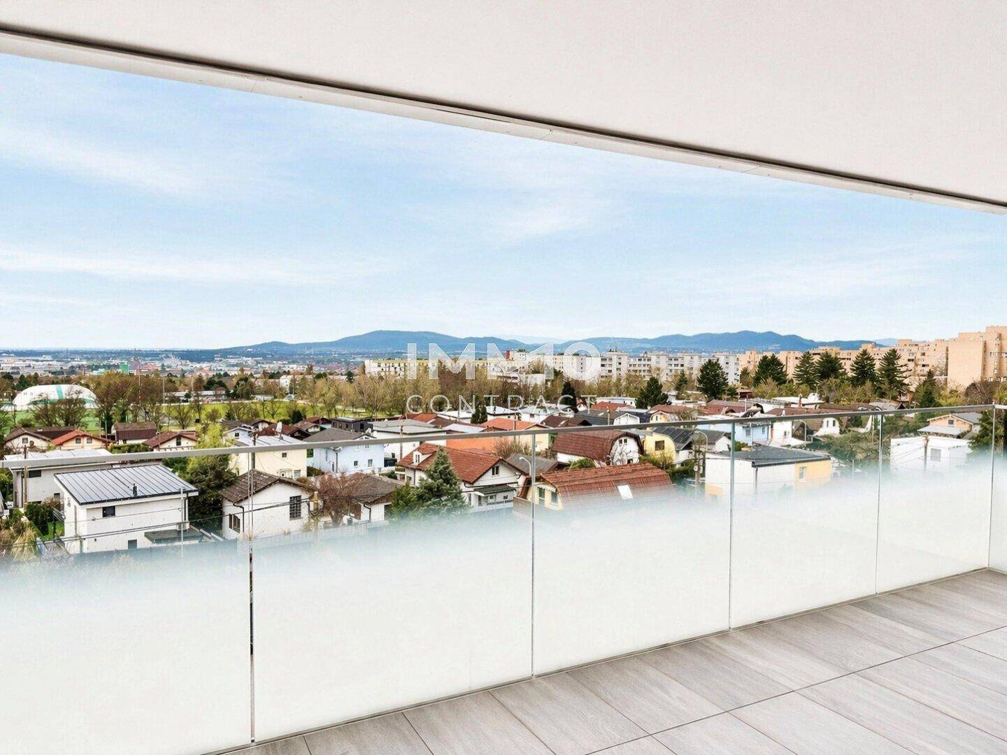 Balkon mit Glasgeländer und weitem Blick über die Stadt und die umliegende grüne Landschaft.