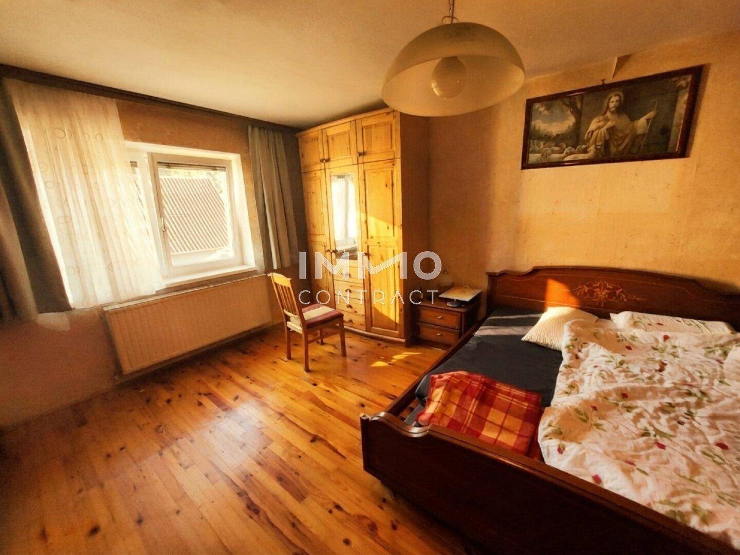 Schlafzimmer mit Holzboden, Holzschrank und einem Bett unter einem Fenster.