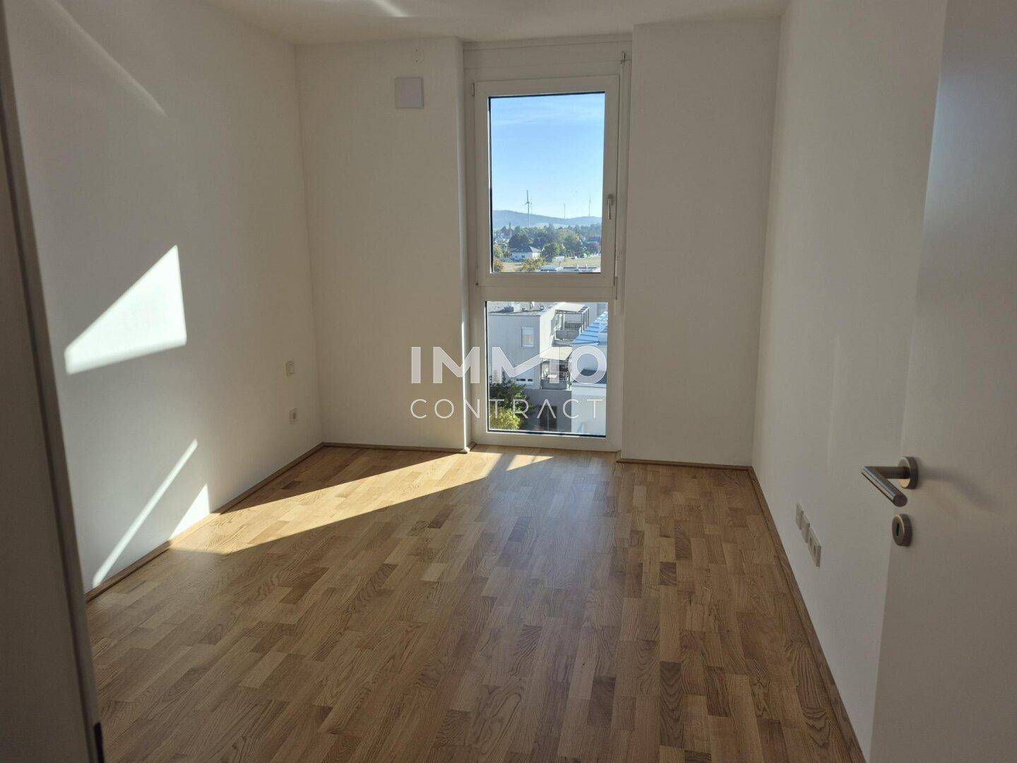 Helles, leeres Zimmer mit Holzparkettboden und großem Fenster, das viel Tageslicht hereinlässt.