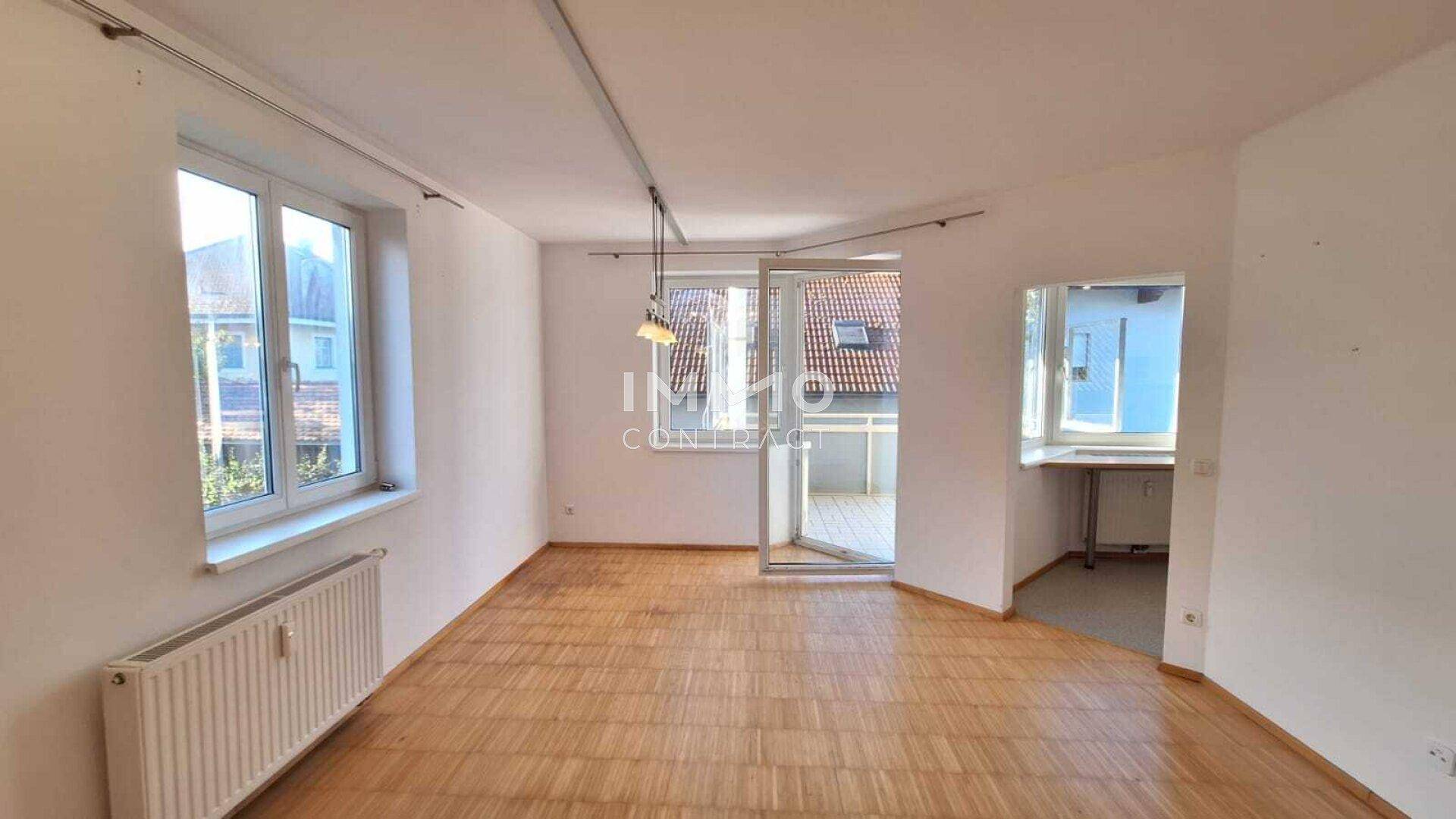 Heller, leerer Wohnraum mit Parkettboden, großen Fenstern und Zugang zu einem Balkon.
